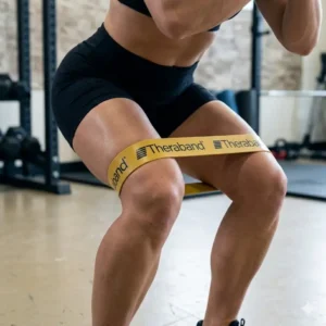 A focused close-up of a muscular female athlete performing a deep squat with a Theraband Gold heavy-duty resistance band stretched taut around her mid-thighs, showing clear muscle definition and significant band tension. The background is a detailed gym environment.